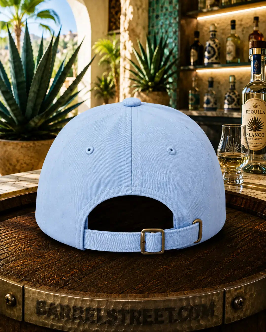 A light blue baseball cap shown from the back, resting on a wooden barrel in a bar setting with a bottle of blanco tequila and a glass nearby.