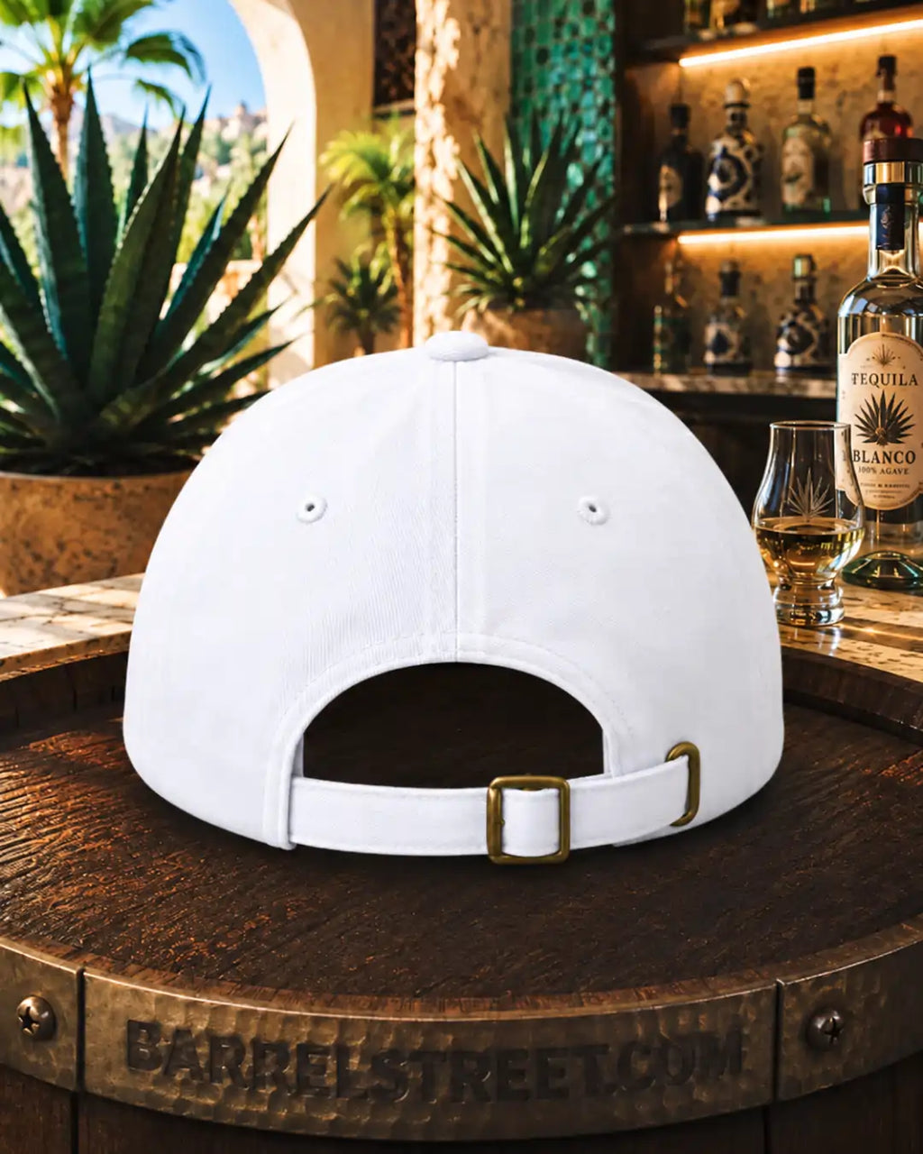 A white baseball cap with an adjustable strap and gold-tone buckle, shown from the back on a rustic wooden barrel in a bar setting with agave plants and liquor bottles in the background.