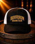 A black and white trucker cap with 'BOURBON HUNTER' embroidered in gold on the front, resting on a wooden barrel with a Barrelstreet.com band visible in the foreground.