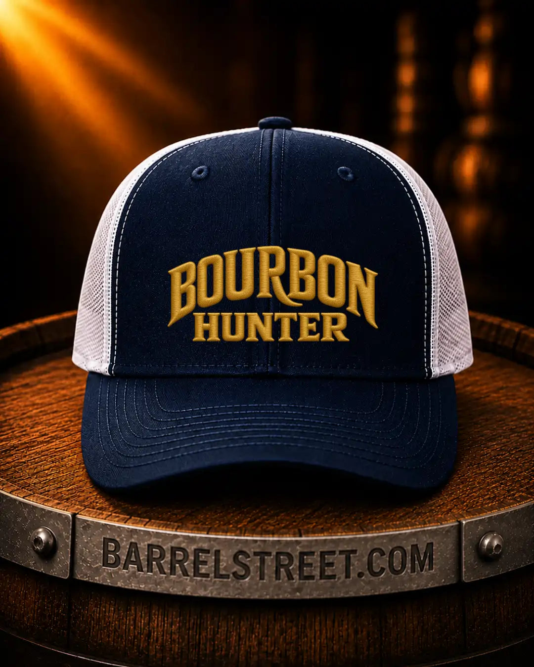A navy blue and white trucker hat with 'BOURBON ENERGY' embroidered in gold on the front, displayed on a wooden barrel with a warm, dark background.