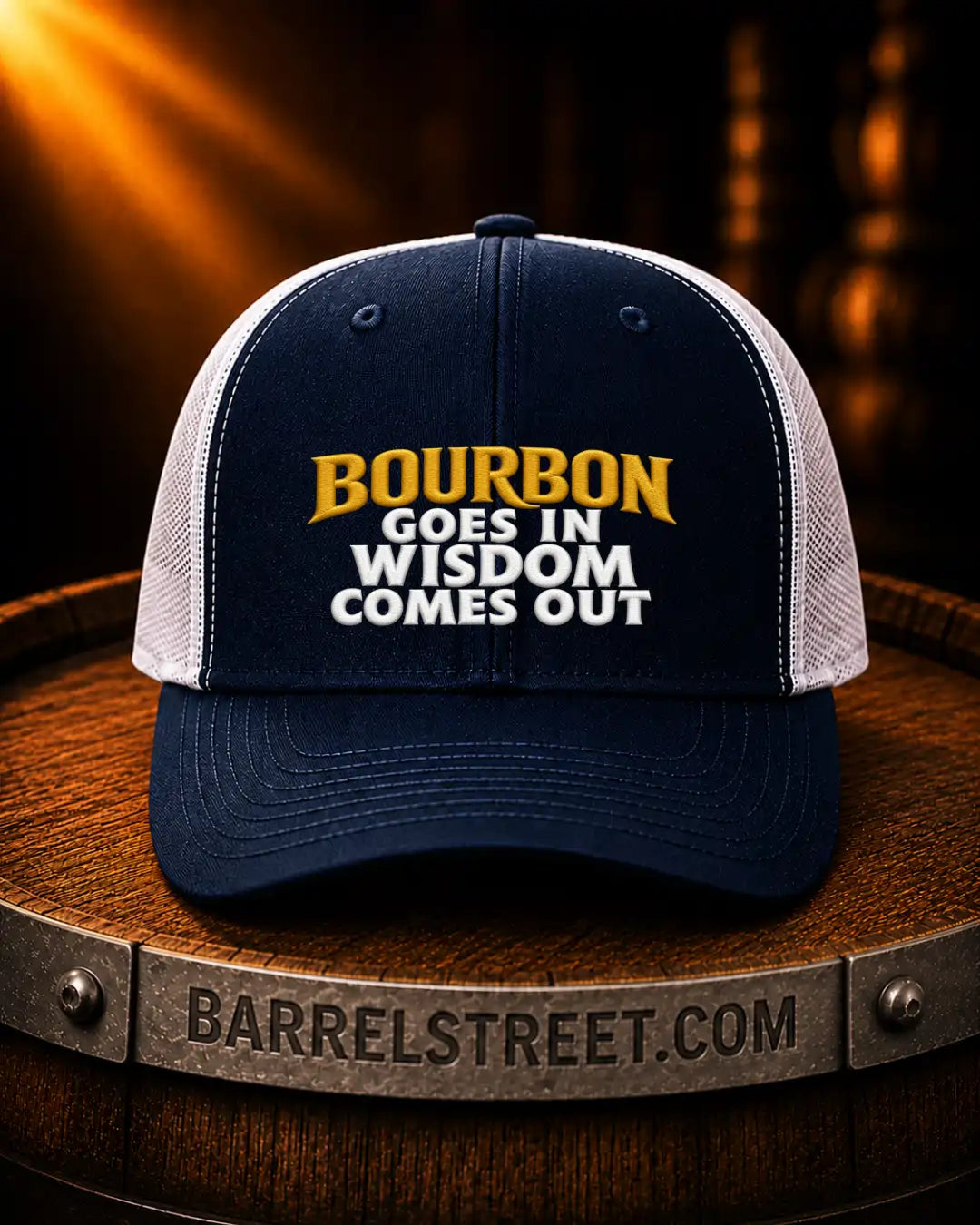 A navy and white trucker cap with the text 'BOURBON GOES IN WISDOM COMES OUT' in yellow and white embroidery, displayed on a wooden barrel with a warm, dramatic background.