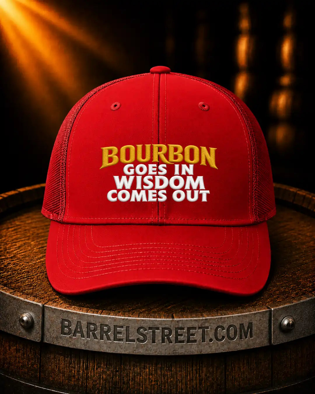 A red trucker-style baseball cap with embroidered text reading 'BOURBON GOES IN WISDOM COMES OUT' in yellow and white, displayed on a wooden barrel. The barrel band shows the text 'BARRELSTREET.