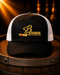 Black and white trucker cap with a gold embroidered Bourbon logo and a gold-and-red bullet graphic on the front, displayed on a wooden barrel with warm spotlighting.