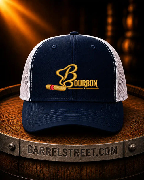 A navy and white trucker cap with a gold embroidered Bourbon logo and a cigar graphic, displayed on a wooden barrel with a Barrelstreet.com banner in the foreground.
