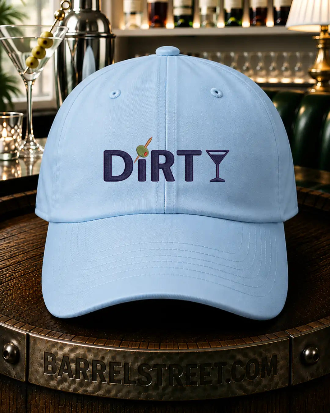 A light blue baseball cap with 'DiRTY' and a martini glass graphic embroidered on the front, displayed on a wooden barrel in a bar setting.