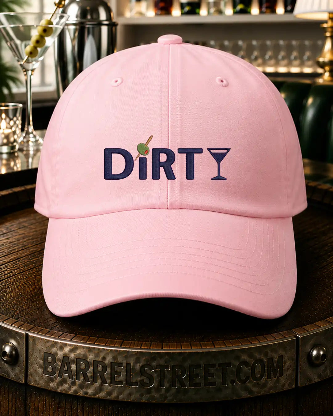 A light pink baseball cap with 'DiRTY' and a martini glass graphic embroidered on the front, resting on a wooden barrel in a bar setting.