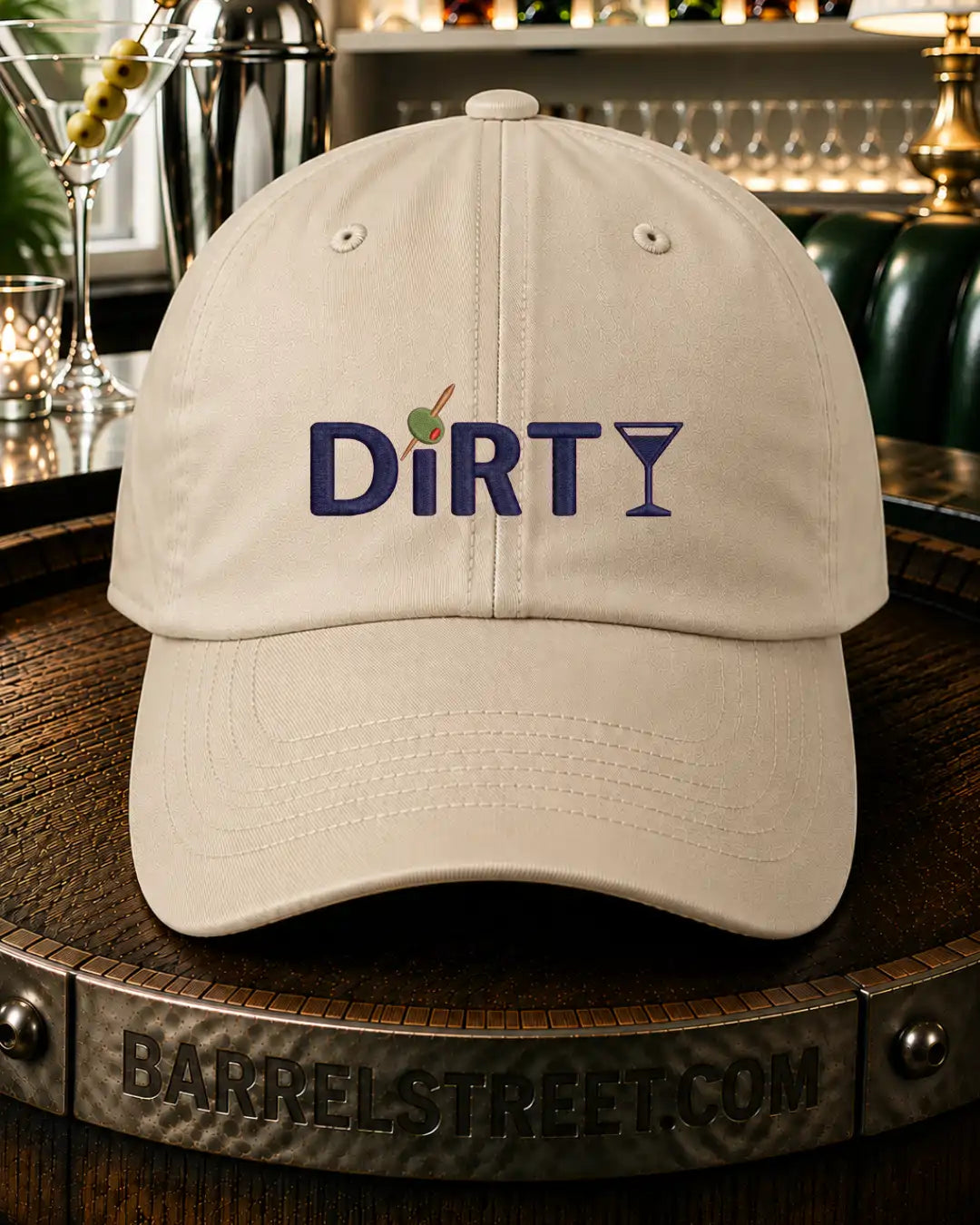 An off-white baseball cap with 'DiRTY' embroidered on the front, featuring a small green olive on a toothpick replacing the letter 'I'. The cap is displayed on a wooden barrel in a bar setting.