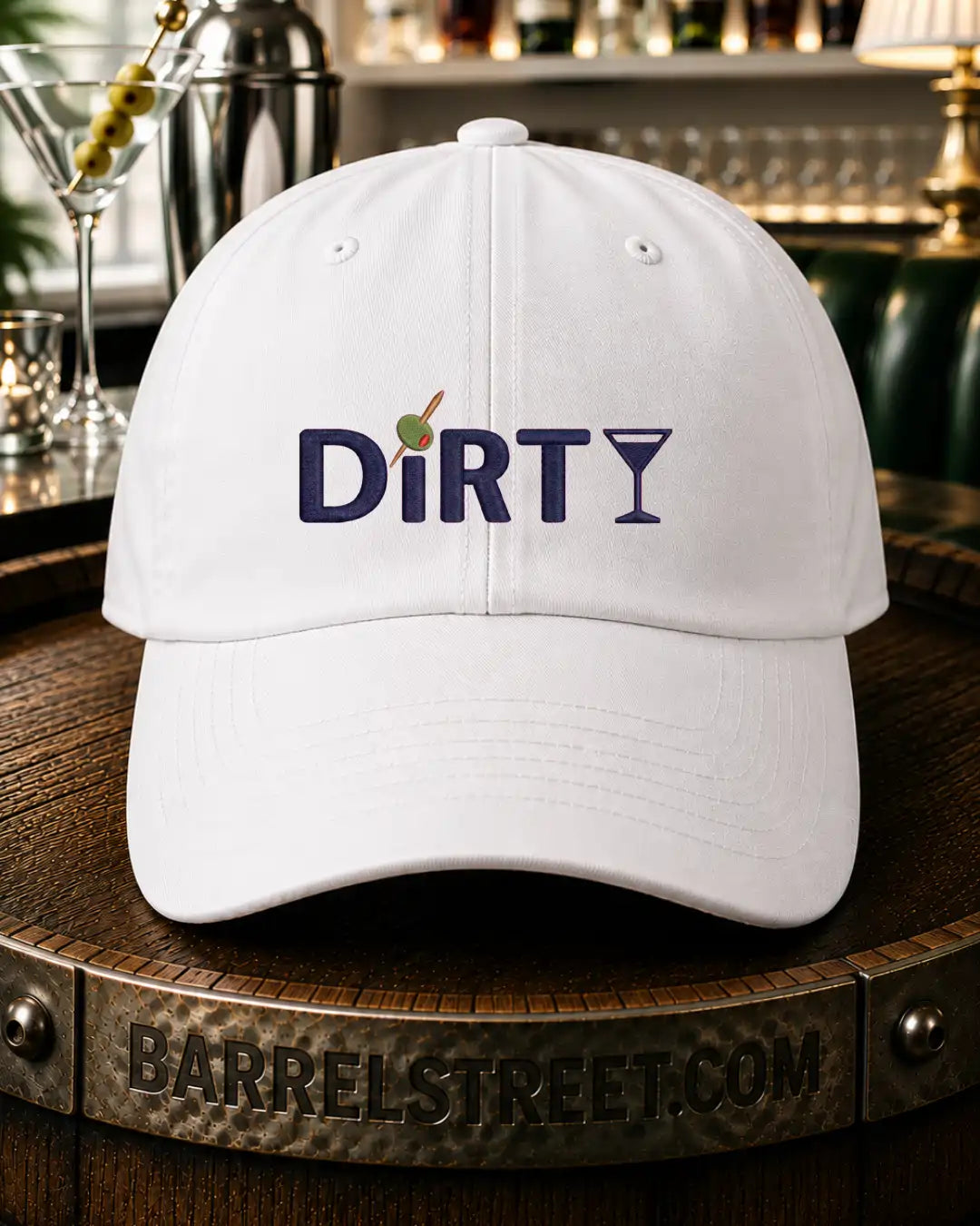 A white baseball cap with 'DiRTY' embroidered on the front, featuring a martini glass icon and a green olive on a toothpick, resting on a wooden barrel in a bar setting.
