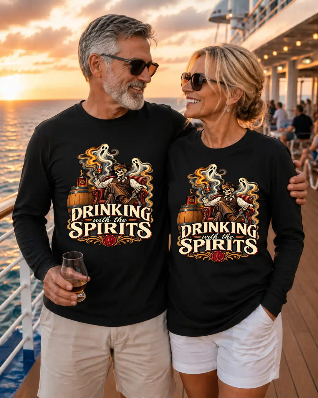 A man and a woman standing together on a cruise ship deck at sunset, both wearing matching black long-sleeve shirts with a graphic print that reads 'DRINKING with the SPIRITS' and showing a ghost, whiskey glass, and barrel illustration.
