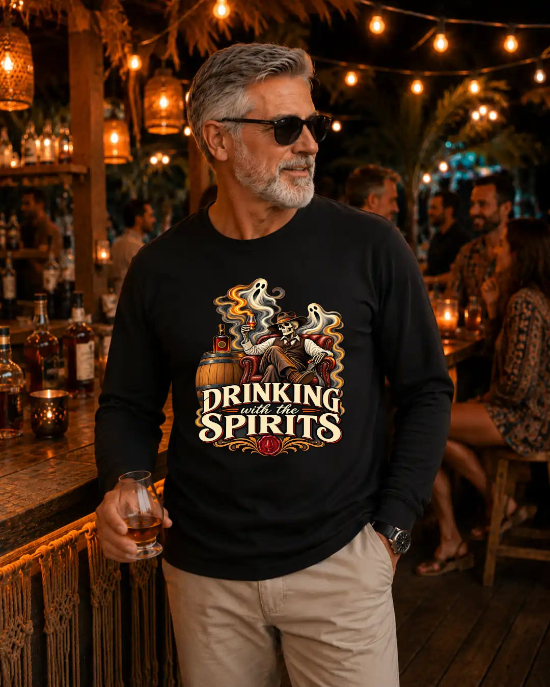 A man wearing a black long-sleeve graphic t-shirt with a 'Drinking with the Spirits' ghost and whiskey illustration, standing in a warmly lit bar environment.