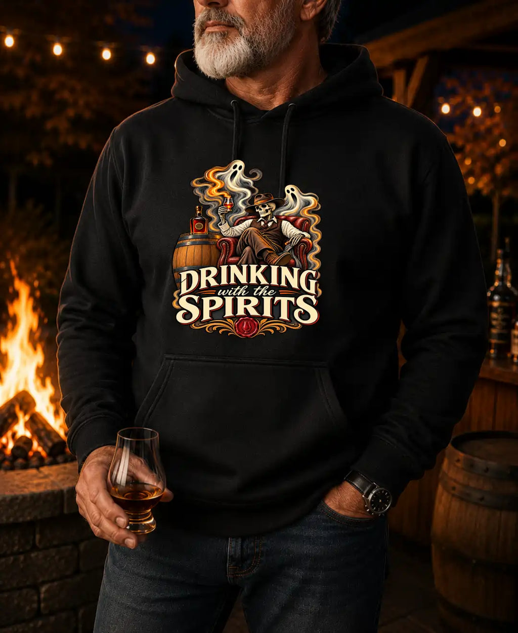 A man wearing a black hoodie with a graphic print that reads 'DRINKING with the SPIRITS' and shows a skeleton at a barrel with a glass, standing in front of a warm fire pit at night.