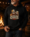 A man wearing a black hoodie with a graphic print that reads 'DRINKING with the SPIRITS' and shows a skeleton at a barrel with a glass, standing in front of a warm fire pit at night.