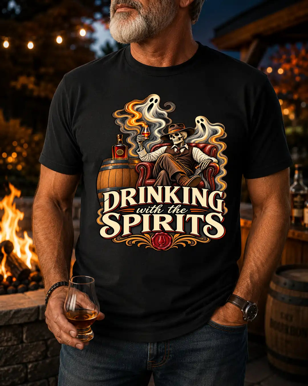 A man wearing a black graphic t-shirt with a Halloween-themed design that reads 'DRINKING with the SPIRITS' and shows a skeleton drinking from a barrel with ghosts and a whiskey glass, set against a warm, outdoor fire pit background.