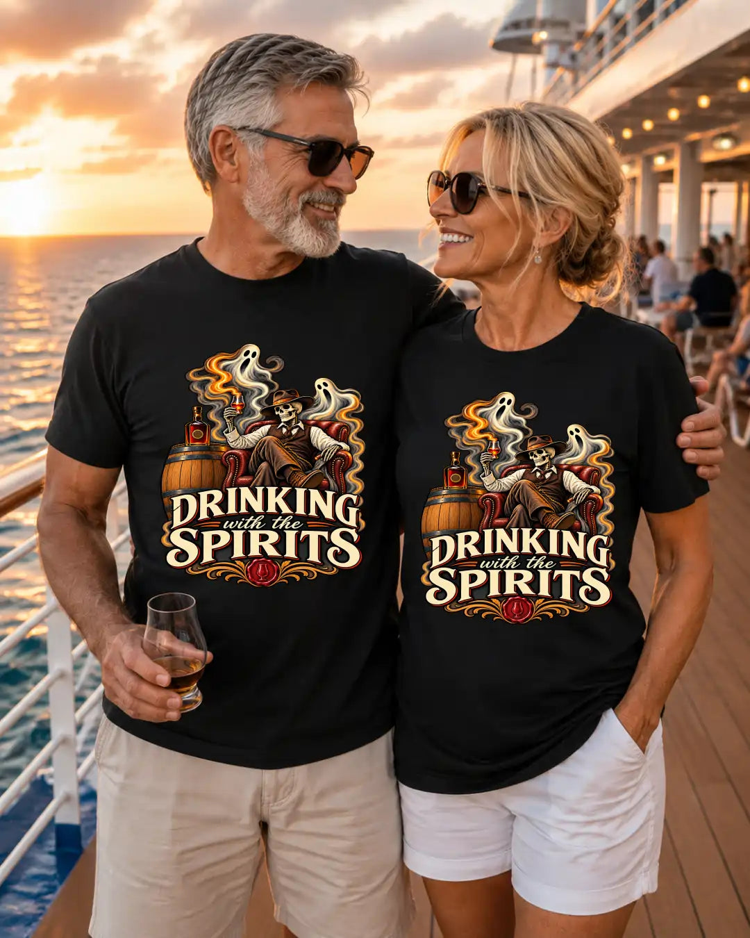 A man and a woman standing together on a cruise ship deck at sunset, both wearing matching black graphic T-shirts with a vintage-style illustration of a skull, whiskey bottle, and the text 'DRINKING with the SPIRITS'.