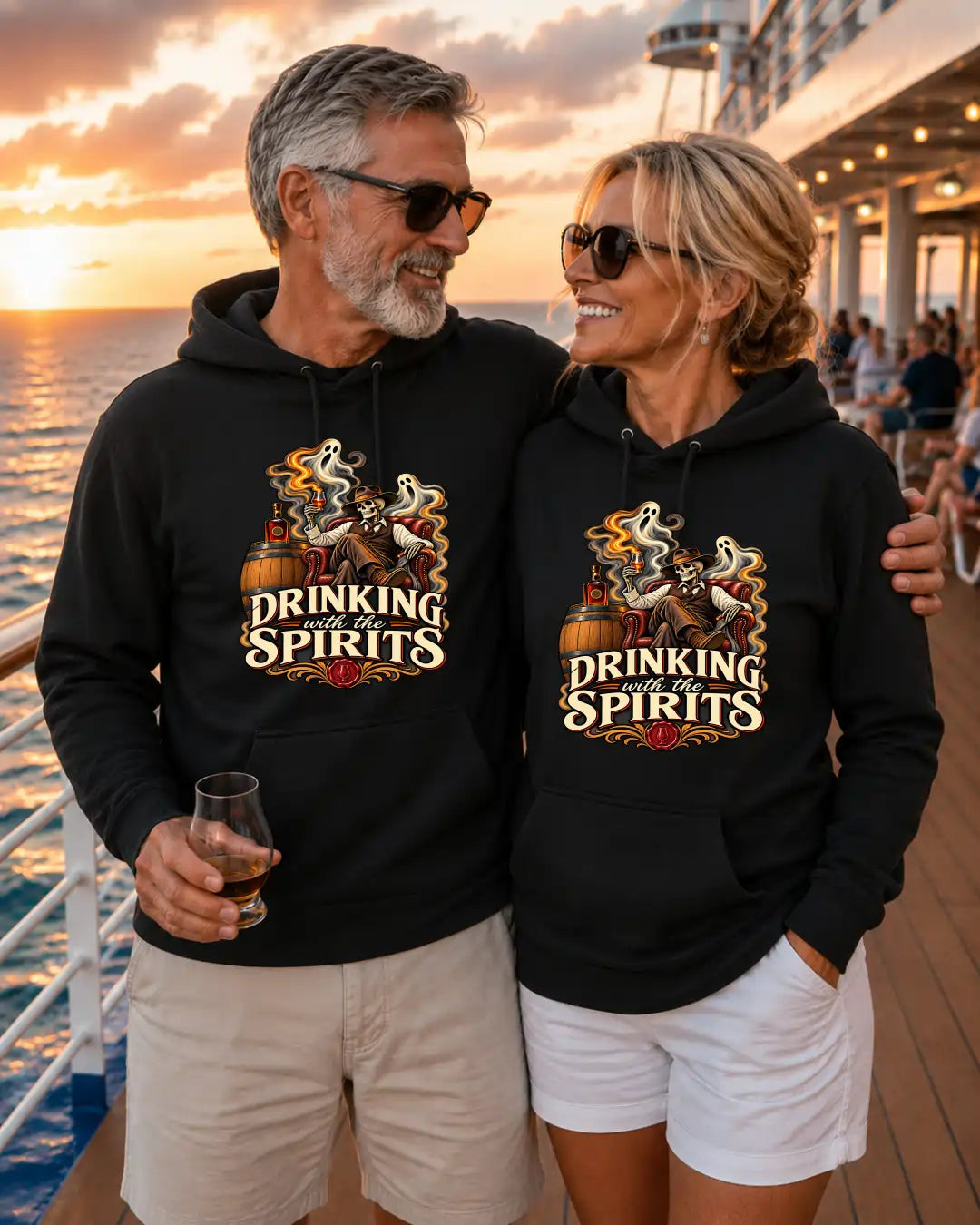 A man and a woman standing together on a cruise ship deck at sunset, both wearing matching black hoodies with a graphic print that reads “DRINKING SPIRITS” and shows a ghost, wine glass, and bottle illustration.