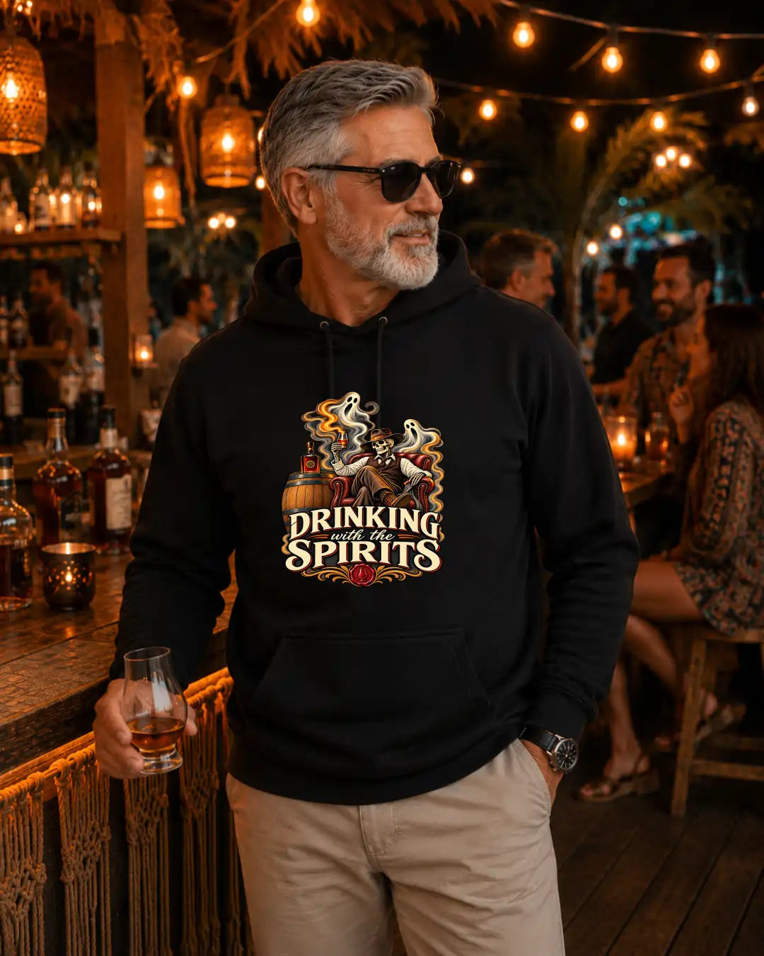 A man wearing a black hoodie with a 'Drinking Spirits' graphic print stands in a warmly lit bar environment, holding a glass of whiskey.