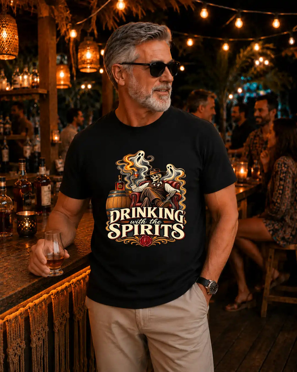 A man wearing a black graphic t-shirt with a 'Drinking with the Spirits' ghost and whiskey design, standing in a warmly lit bar environment.