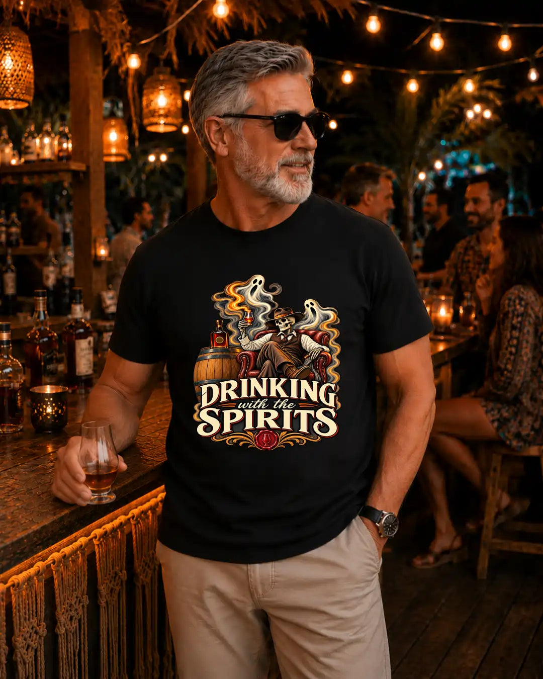 A man wearing a black graphic t-shirt with a 'Drinking with the Spirits' ghost and whiskey design, standing in a warmly lit bar environment.