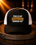 A black and white trucker hat with the text 'THIS IS MY BOURBON DRINKING HAT' on the front, displayed on a wooden barrel. The barrel has 'BARRELSTREET.