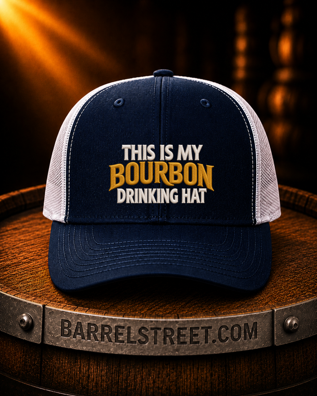 A navy blue and white trucker hat with the text 'THIS IS MY BOURBON DRINKING HAT' embroidered on the front, displayed on a wooden barrel with a warm, glowing background.