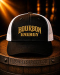 A black and white trucker hat with 'BOURBON ENERGY' embroidered in gold on the front, displayed on a wooden barrel with a dark, warm background.