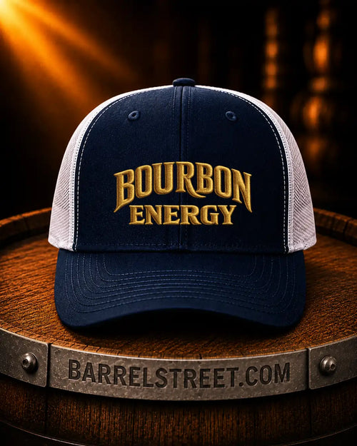 A navy blue and white trucker hat with 'BOURBON ENERGY' embroidered in gold on the front, displayed on a wooden barrel with a warm, dark background.