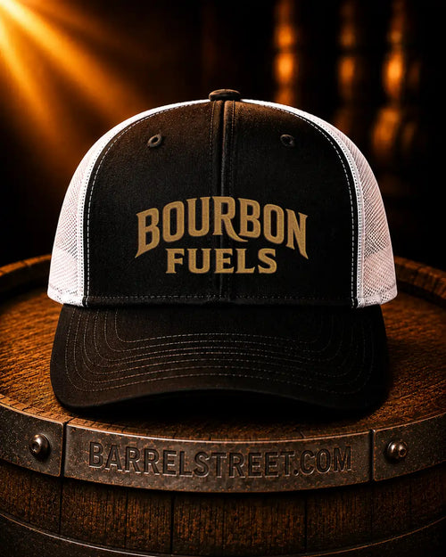 A black and white trucker hat with 'BOURBON FUELS' embroidered in gold on the front, resting on a wooden barrel with a Barrel Street branding band visible.