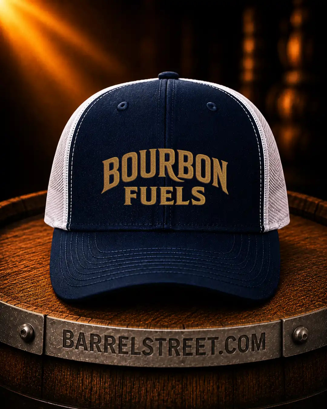 A navy blue and white trucker hat with 'BOURBON FUELS' embroidered in gold on the front, displayed on a wooden barrel with a Barrelstreet.com banner in the foreground.