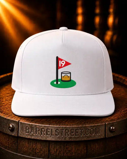 A white structured baseball cap with a small graphic of a golf hole and a whiskey glass on the front, displayed on a wooden barrel with warm golden light beams in the background. The barrel band reads BARRELSTREET.