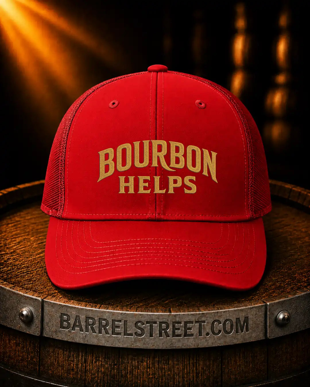 A red trucker-style baseball cap with gold embroidered text reading 'BOURBON HELPS' sits on a wooden barrel. The barrel has a metal band engraved with 'BARRELSTREET.