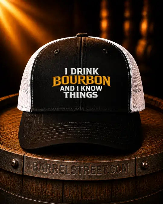 A black and white trucker hat with the text 'I DRINK BOURBON AND I KNOW THINGS' embroidered on the front, displayed on a wooden barrel with a warm, dark background.