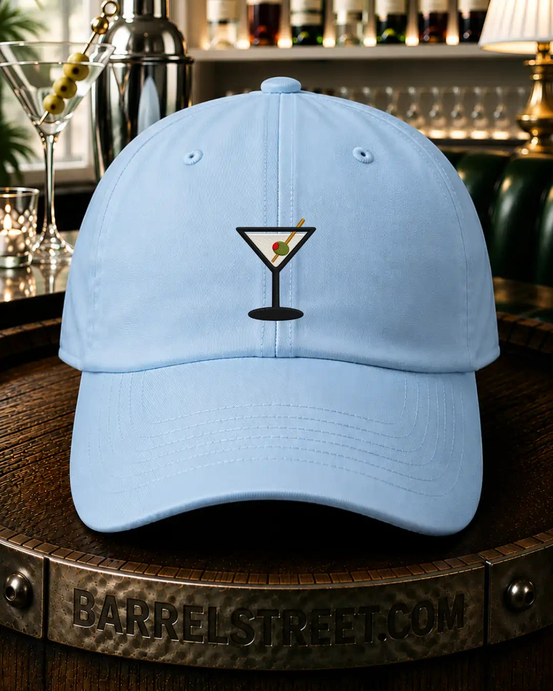 A light blue baseball cap featuring a black martini glass graphic with a green olive on the front, displayed on a wooden barrel with a bar scene in the background.