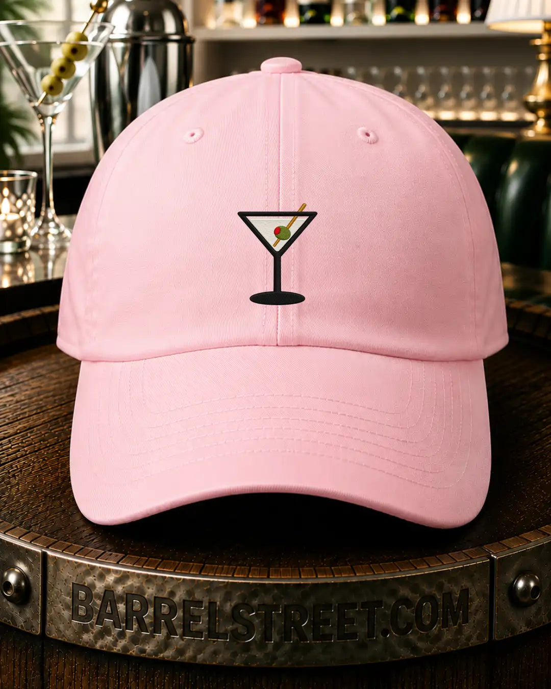 A light pink baseball cap with a black martini glass graphic on the front, resting on a wooden barrel with a bar scene blurred in the background.