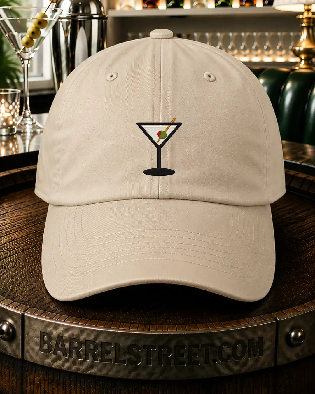 A light beige baseball cap with a black martini glass graphic on the front, displayed on a wooden barrel in a bar setting.