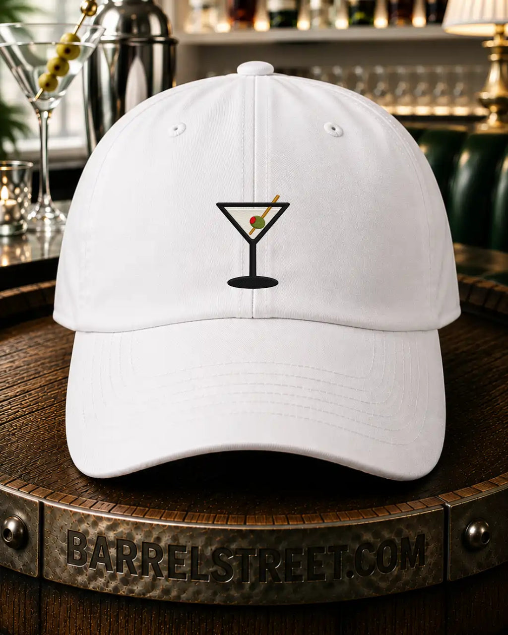 A white baseball cap with a black martini glass graphic on the front, featuring a red and green olive detail, displayed on a wooden barrel with a bar background.
