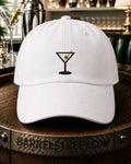 A white baseball cap with a black martini glass graphic on the front, featuring a red and green olive detail, displayed on a wooden barrel with a bar background.