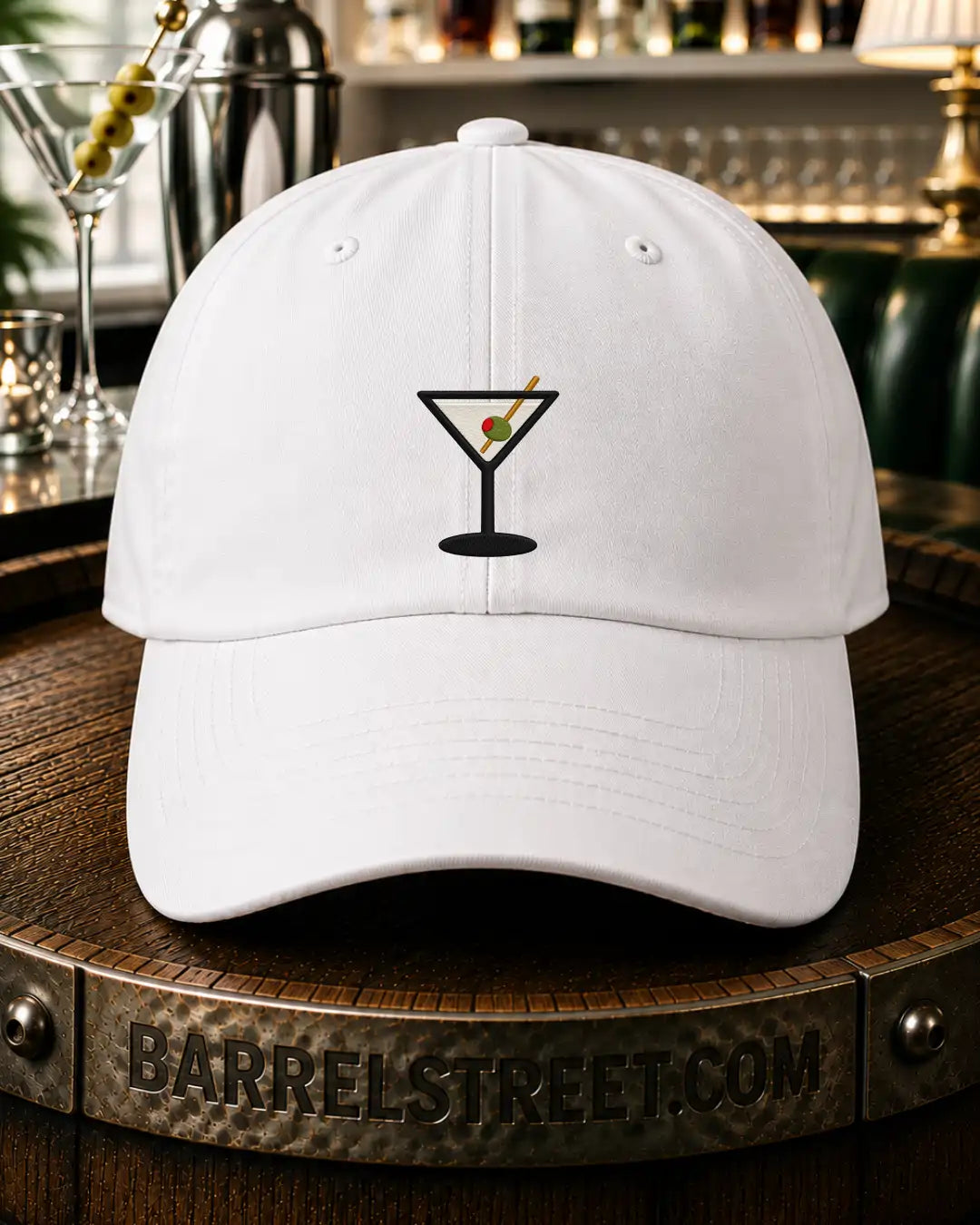 A white baseball cap with a black martini glass graphic on the front, featuring a red and green olive detail, displayed on a wooden barrel with a bar background.