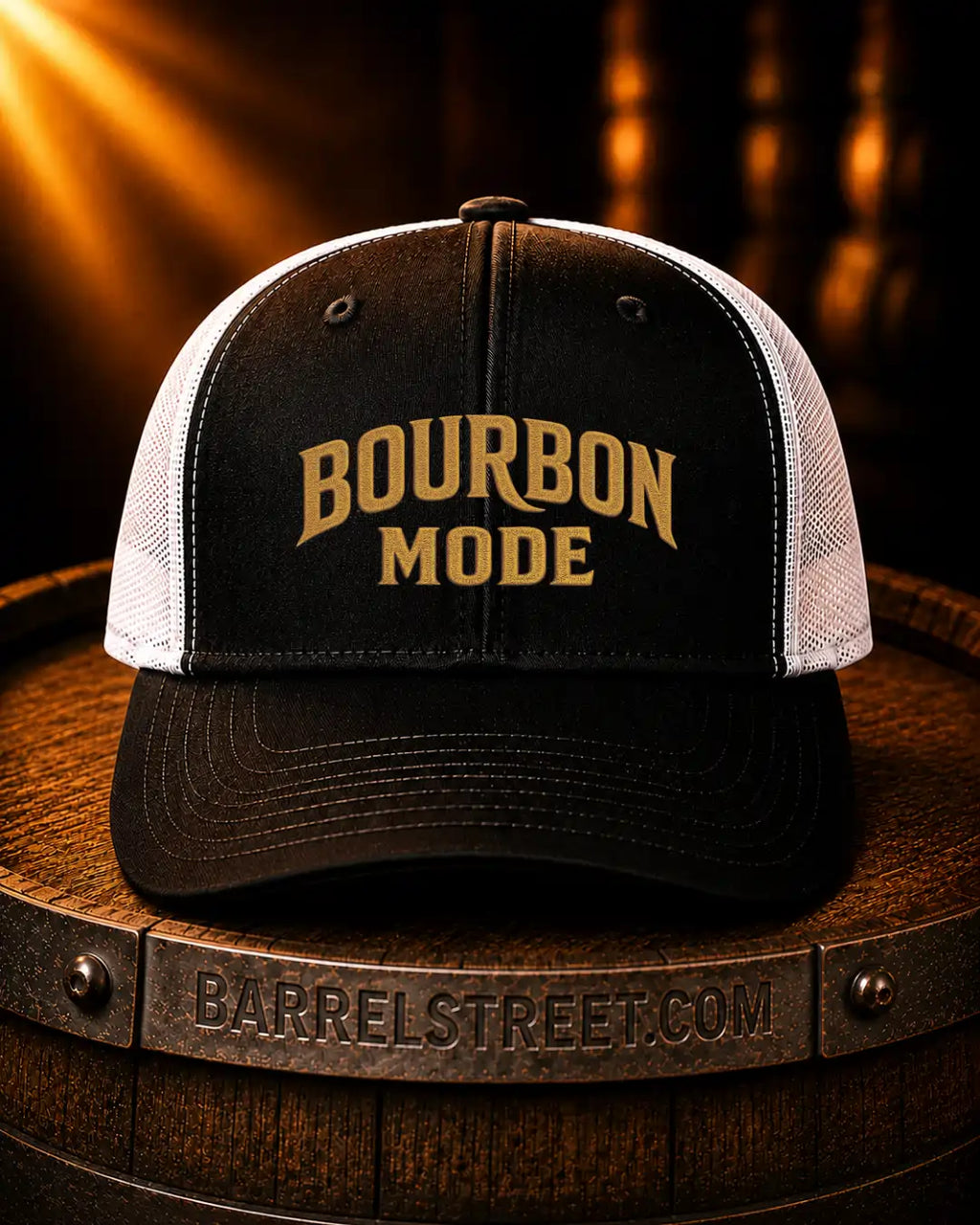 A black and white trucker hat with 'BOURBON MODE' embroidered in gold on the front, displayed on a wooden barrel with warm spotlighting.