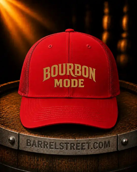 A red trucker-style cap with 'BOURBON MODE' embroidered in gold on the front, displayed on a wooden barrel with a metal band reading 'BARRELSTREET.COM'.