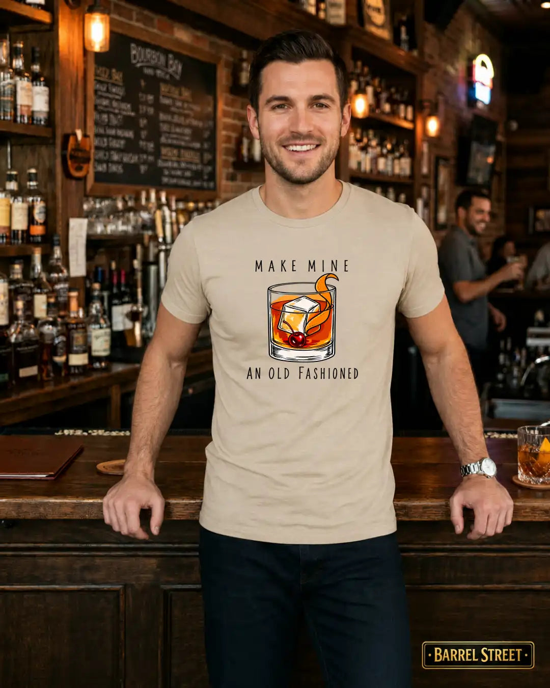 A man standing behind a wooden bar in a barbershop-style interior, wearing a beige graphic t-shirt with a printed illustration of a whiskey glass and the text 'MAKE MINE AN OLD FASHIONED'.
