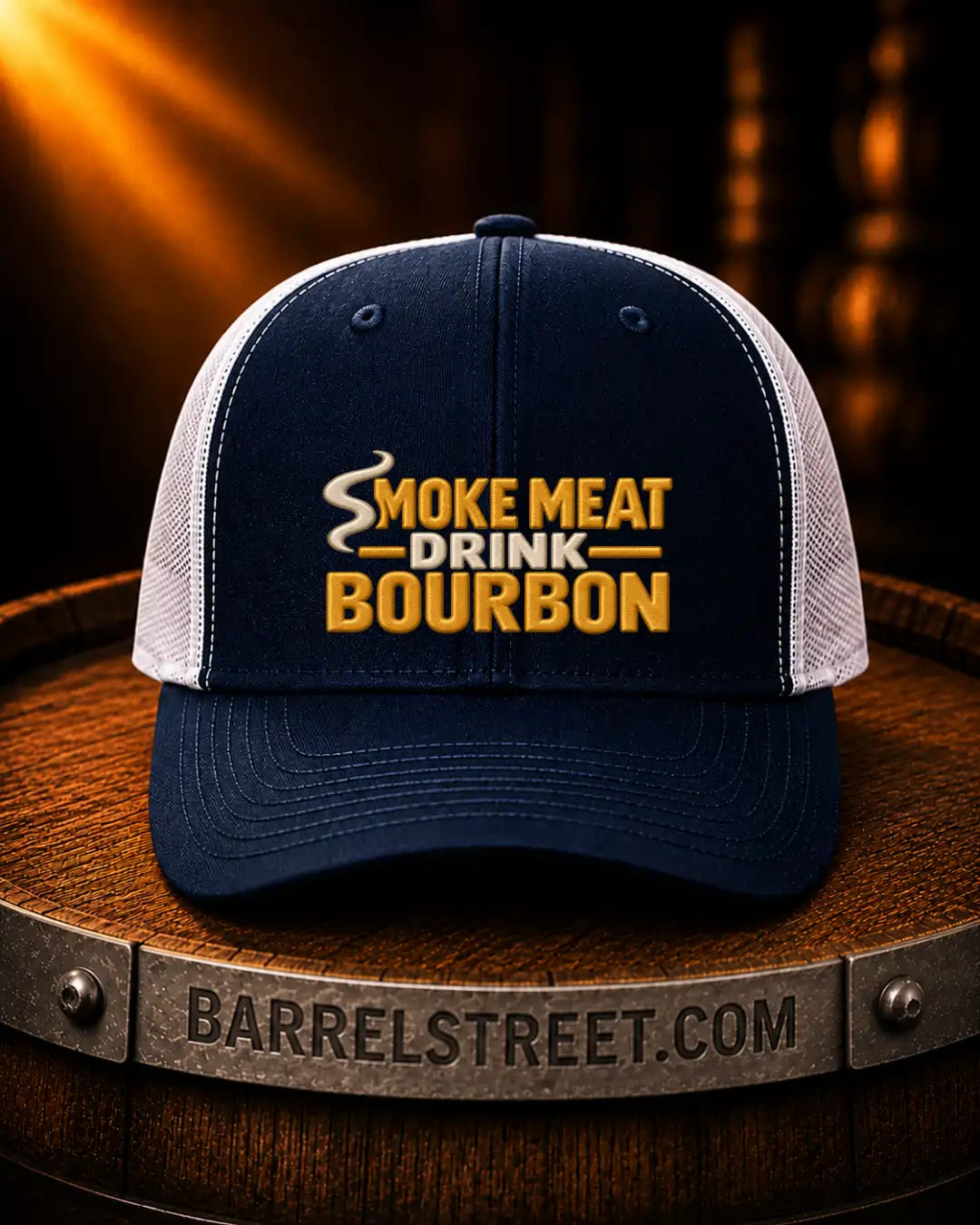 A navy blue and white trucker hat with yellow embroidered text reading 'SMOKE MEAT DRINK BOURBON' sits on a wooden barrel. The barrel band shows the text 'BARRELSTREET.