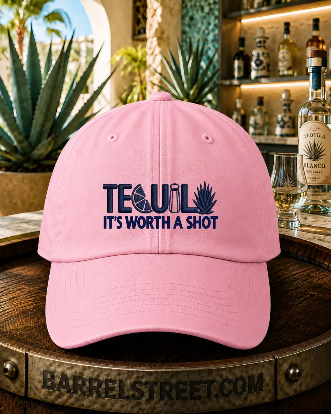 A light pink baseball cap with navy blue embroidered text reading 'TEQUIL IT'S WORTH A SHOT' sits on a wooden barrel in a bar setting with bottles and agave plants in the background.