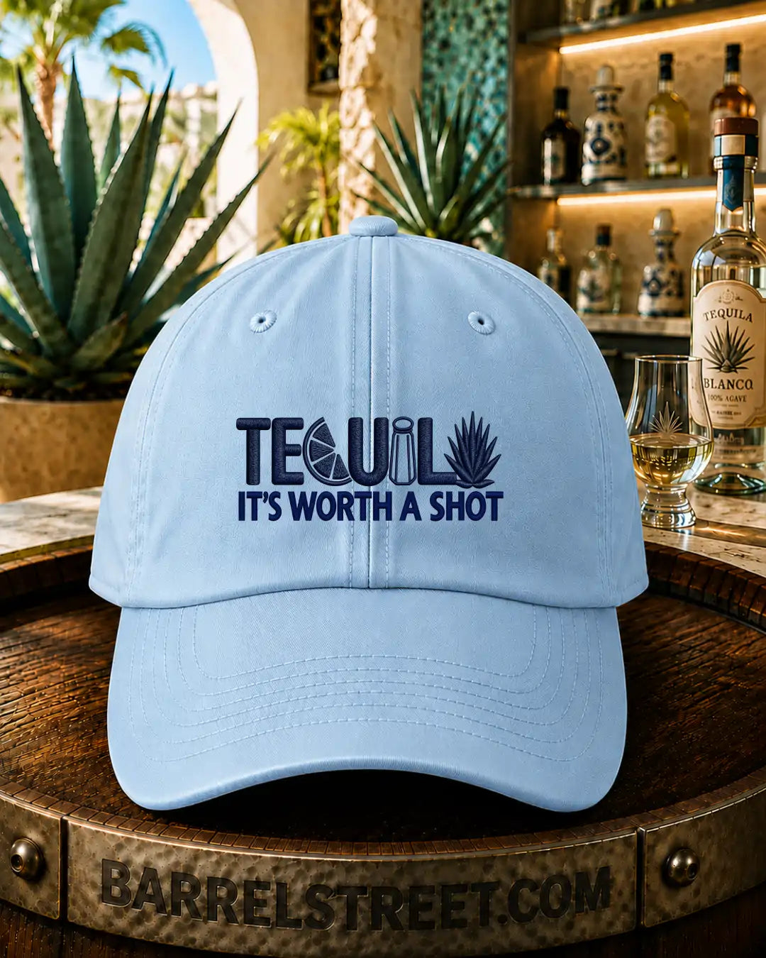A light blue baseball cap with dark blue embroidered text reading 'TEQUIL It's worth a shot' sits on a wooden barrel in a bar setting with a tequila bottle and glass in the background.