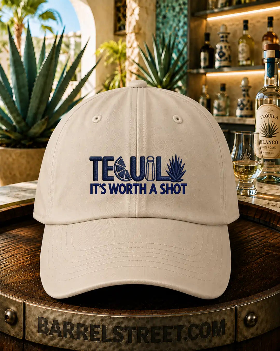 A light beige baseball cap with a dark blue embroidered graphic reading 'TEQUIL IT'S WORTH A SHOT' sits on a wooden barrel in a bar setting with tequila bottles and a glass of tequila in the background.