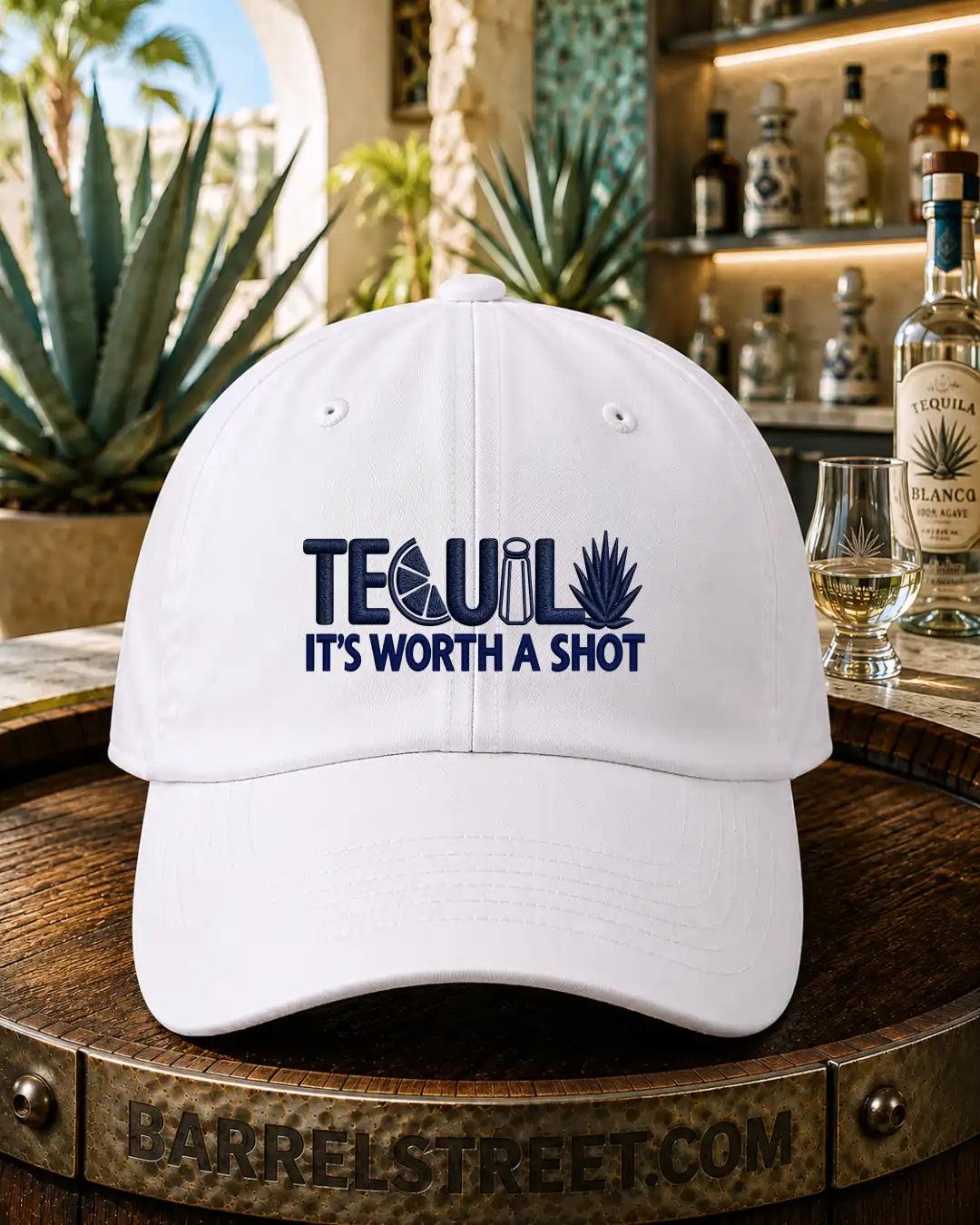 A white baseball cap with navy blue embroidered text reading 'TEQUILA IT'S WORTH A SHOT' sits on a wooden barrel in a bar setting, with bottles, a glass, and plants in the background.