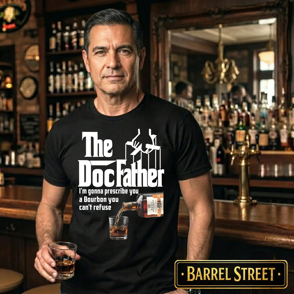 Man wearing a black t-shirt with 'The Doc Father' design in a bar setting, holding a glass of whiskey.