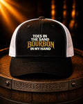 A black and white trucker hat with embroidered text reading 'TOES IN THE SAND BOURBON IN MY HAND' sits on a wooden barrel. The hat has a black front panel and brim, white mesh back panels, and gold and white embroidery.