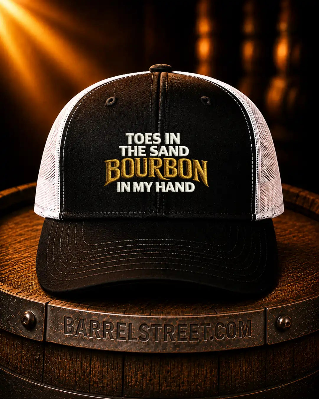 A black and white trucker hat with embroidered text reading 'TOES IN THE SAND BOURBON IN MY HAND' sits on a wooden barrel. The hat has a black front panel and brim, white mesh back panels, and gold and white embroidery.