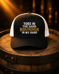 A black and white trucker hat with the text 'TOES IN THE SAND BOURBON IN MY HAND' in white and gold lettering, displayed on a wooden barrel.