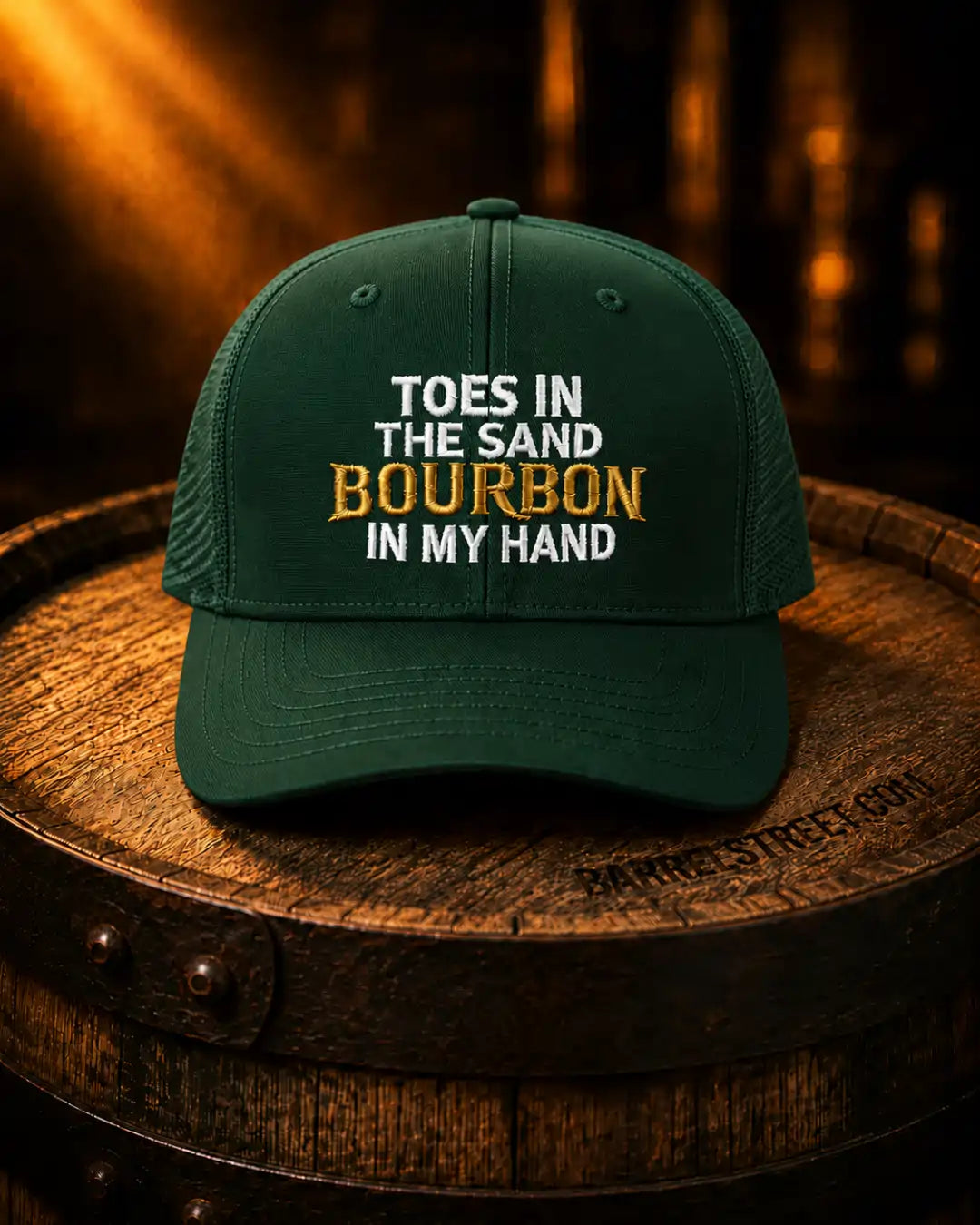 A dark green trucker-style baseball cap with embroidered text reading 'TOES IN THE SAND BOURBON IN MY HAND' in white and gold, displayed on top of a wooden barrel in a warm, dimly lit setting.